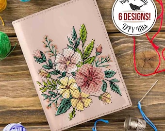 Op de afbeelding: Een roze DIY-kit met een bloemenkruissteekontwerp. De kit bevat een naald, vingerhoed en draad. De omslag toont een kruisgestikt boeket van roze en gele bloemen met groene bladeren. De tekst luidt "high quality" en "6 designs! DIY Kits."
