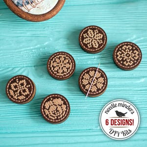 Könnte beinhalten: Sechs runde Nadelhalter mit einem braunen Holzrahmen und einem beige-braunen Stickmuster. Eine Nadel liegt auf einem der Halter. Das Bild enthält auch einen Aufkleber mit dem Text "needle minders 6 DESIGNS! DIY Kits".