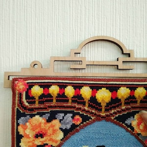 Op de afbeelding: Een kleurrijk geborduurd wandkleed met een bloemenmotief, ingelijst in een houten hanger met een decoratieve boog.