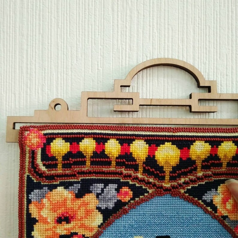 Op de afbeelding: Een kleurrijk geborduurd wandkleed met een bloemenmotief, ingelijst in een houten hanger met een decoratieve boog.