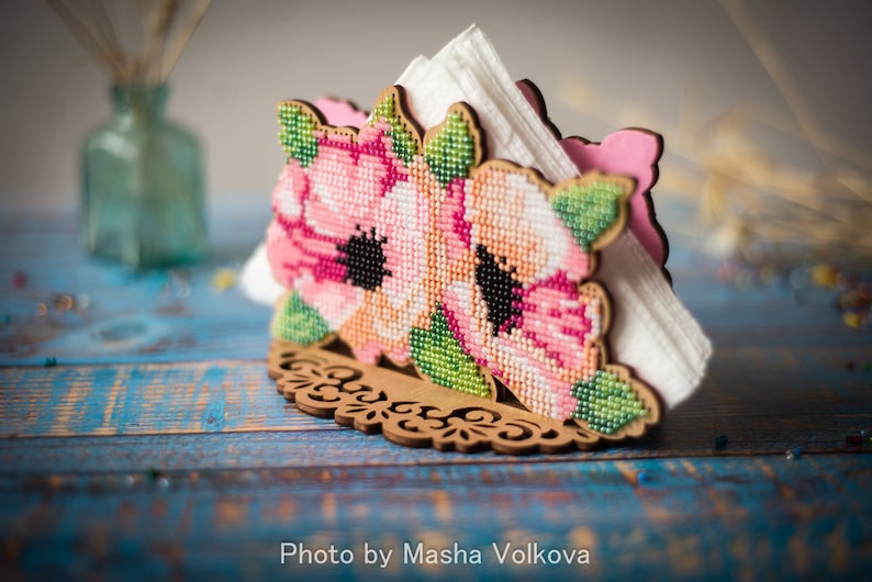 May include: A wooden napkin holder with a floral design. The holder is decorated with pink and green beaded flowers. The holder is sitting on a blue wooden surface.