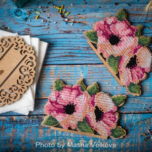 May include: Two wooden napkin holders with a floral design. Each holder features pink and white flowers with green leaves. The flowers are made of beads and are stitched together. The holders are on a blue wooden surface.