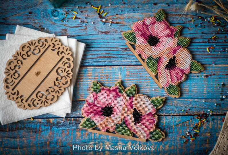May include: Two wooden napkin holders with a floral design. Each holder features pink and white flowers with green leaves. The flowers are made of beads and are stitched together. The holders are on a blue wooden surface.