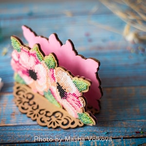 May include: A pink and brown wooden napkin holder with a floral design. The holder is decorated with pink and white flowers and green leaves. The holder is on a blue wooden surface.