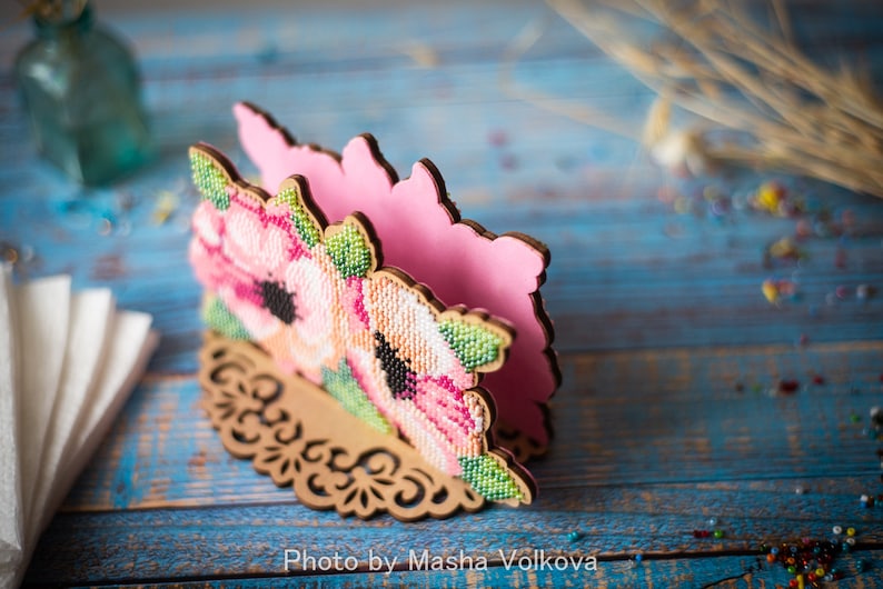 May include: A pink and brown wooden napkin holder with a floral design. The holder is decorated with pink and white flowers and green leaves. The holder is on a blue wooden surface.