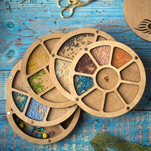 May include: A wooden bead storage organizer with multiple rotating compartments. Each compartment is filled with colorful beads. The organizer is on a blue wooden surface.