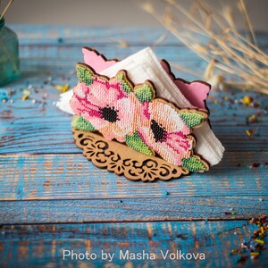 May include: A wooden napkin holder with a floral design. The holder is decorated with pink and white flowers and green leaves. The holder is sitting on a blue wooden table.