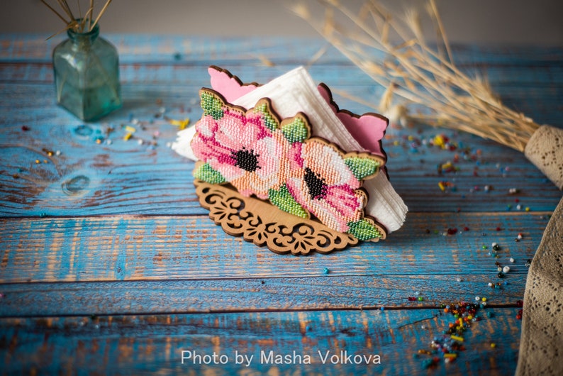 May include: A wooden napkin holder with a floral design. The holder is decorated with pink and white flowers and green leaves. The holder is sitting on a blue wooden table.