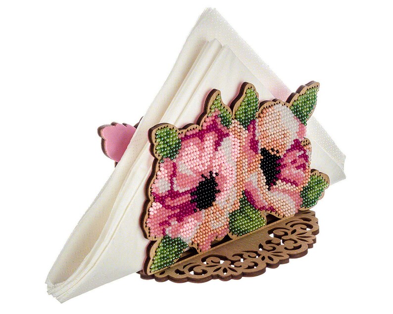 May include: A wooden napkin holder with a floral design. The holder is decorated with pink and green beads, creating a floral pattern. The holder is designed to hold a napkin in a triangular shape.