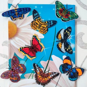 May include: A collection of eight beaded butterfly ornaments in various colors and patterns, including blue, red, yellow, orange, black, and white. The butterflies are arranged on a blue and white background with a large white daisy.