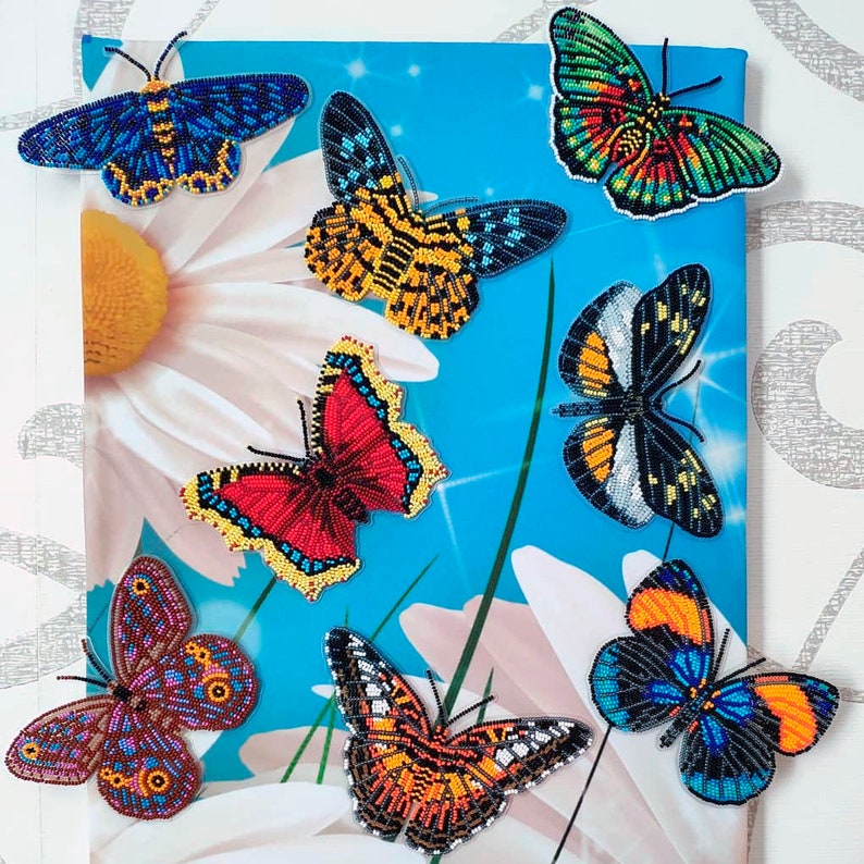 May include: A collection of eight beaded butterfly ornaments in various colors and patterns, including blue, red, yellow, orange, black, and white. The butterflies are arranged on a blue and white background with a large white daisy.