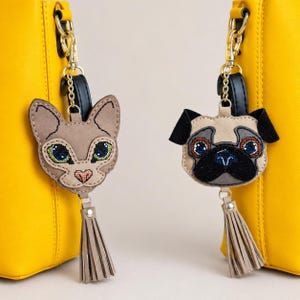 May include: Two bag charms attached to yellow bags. One charm is a tan cat head with green eyes and a pink nose, the other is a tan pug head with black ears and a black muzzle. Both charms feature embroidered details and a beige leather tassel.