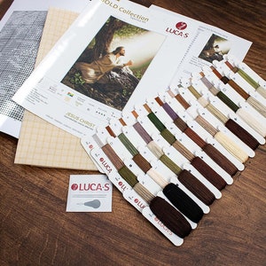 May include: A cross-stitch kit featuring a depiction of Jesus Christ. The kit includes a printed pattern, Aida cloth, and a selection of DMC embroidery floss in various shades of brown, green, and beige. The kit is labelled "Jesus Christ" and "Gold Collection".