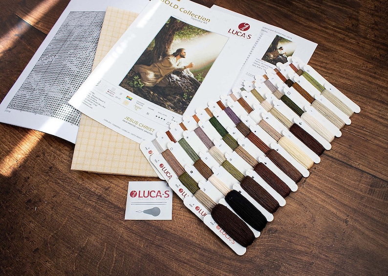 May include: A cross-stitch kit featuring a depiction of Jesus Christ. The kit includes a printed pattern, Aida cloth, and a selection of DMC embroidery floss in various shades of brown, green, and beige. The kit is labelled "Jesus Christ" and "Gold Collection".