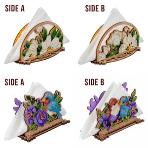 May include: Two wooden napkin holders with different designs. The first holder features a white floral design with green leaves. The second holder features two blue birds with purple flowers. Both holders have a white napkin inside.