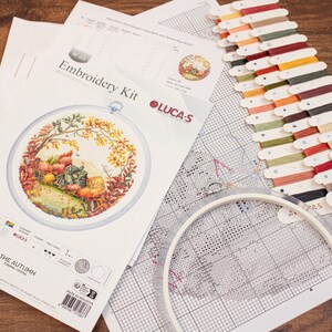 May include: Cross stitch embroidery kit with a colorful autumn scene, including a small house, trees, and leaves. The kit includes a printed fabric, embroidery floss, a needle, and a hoop. The kit is labeled "The Autumn" and "Lucas".