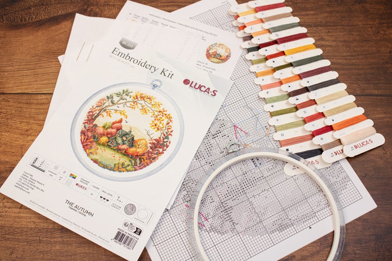May include: Cross stitch embroidery kit with a colorful autumn scene, including a small house, trees, and leaves. The kit includes a printed fabric, embroidery floss, a needle, and a hoop. The kit is labeled "The Autumn" and "Lucas".