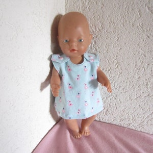 Könnte beinhalten: Eine Puppe in einem hellblauen Kleid mit einem Muster aus rosa und weißen Vögeln. Das Kleid hat Flügelärmel und Knöpfe an den Schultern. Die Puppe steht auf einer rosa Oberfläche.