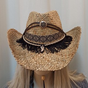 May include: A straw cowboy hat with a brown leather band and a silver sun design. The hat has a black fringe trim and a silver charm.
