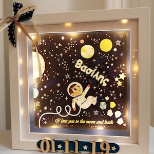 May include: A white light-up picture frame with a black and white illustration of an astronaut in space. The frame has a blue border and the text "Baoiang" and "I love you to the moon and back". The date "01*11*19" is displayed on the bottom of the frame.