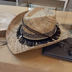 May include: A wide-brimmed straw hat with a black fringe trim and a brown leather band with a geometric pattern. The hat has a silver bead accent.