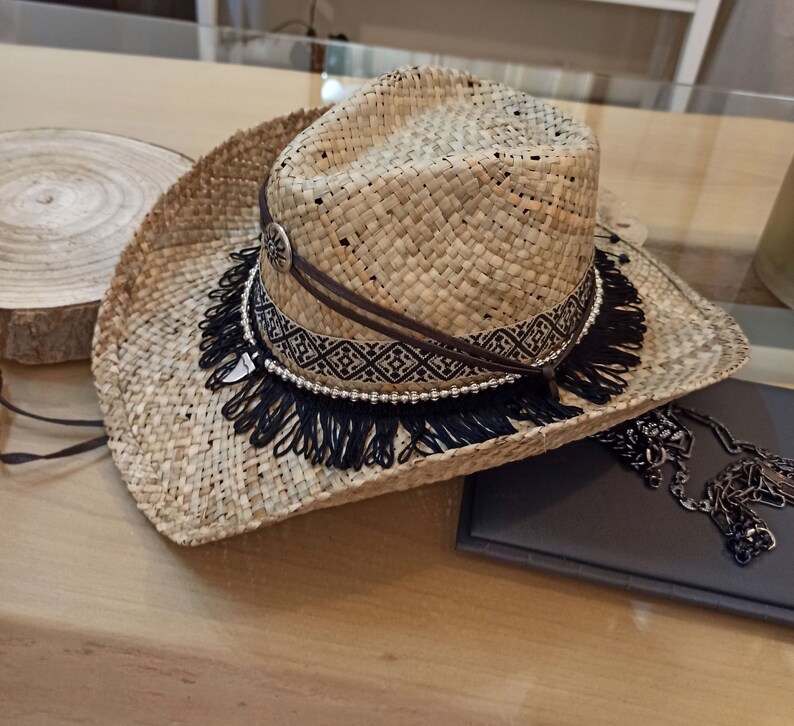 May include: A wide-brimmed straw hat with a black fringe trim and a brown leather band with a geometric pattern. The hat has a silver bead accent.