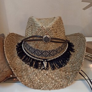 May include: A straw cowboy hat with a black and white beaded band and a black fringe. The hat has a brown leather band with a silver sun design.