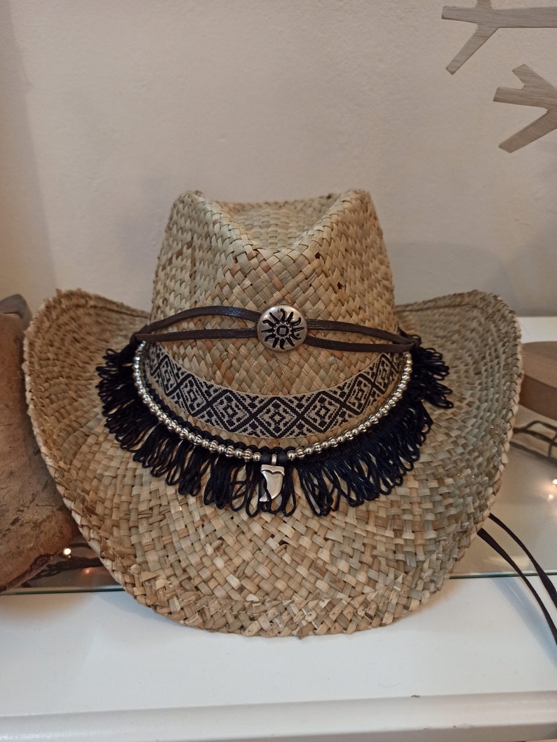 May include: A straw cowboy hat with a black and white beaded band and a black fringe. The hat has a brown leather band with a silver sun design.