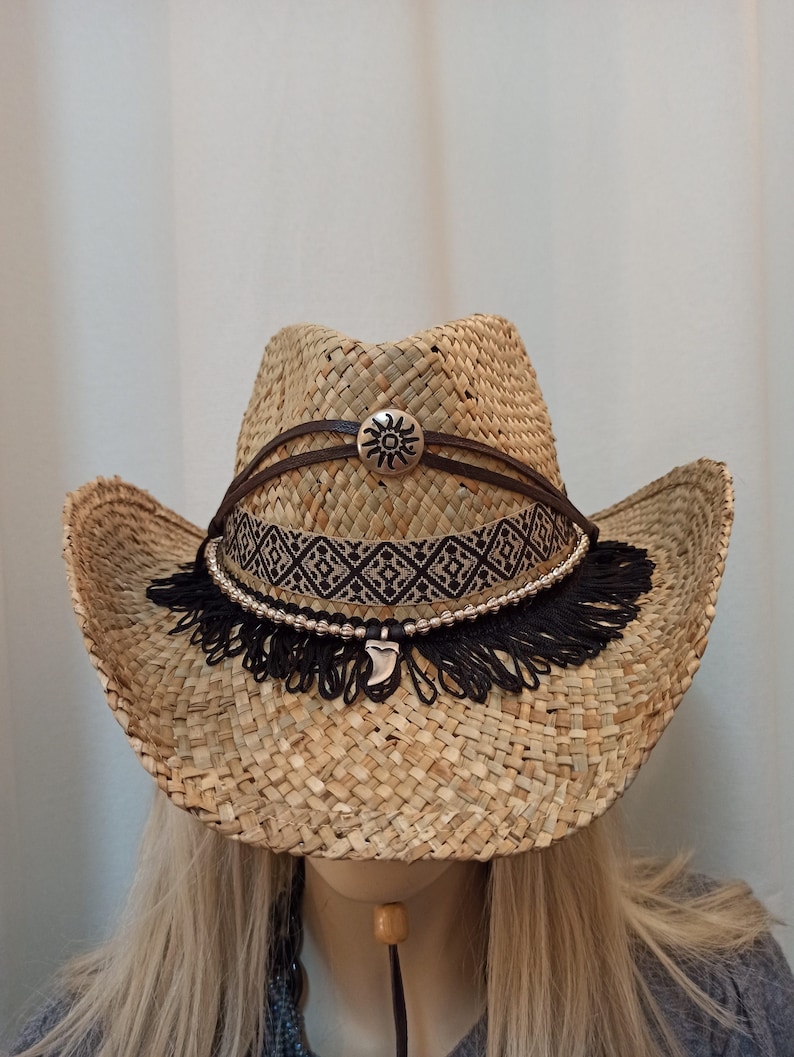 May include: A straw cowboy hat with a brown leather band and a silver sun design. The hat has a black fringe trim and a silver charm.