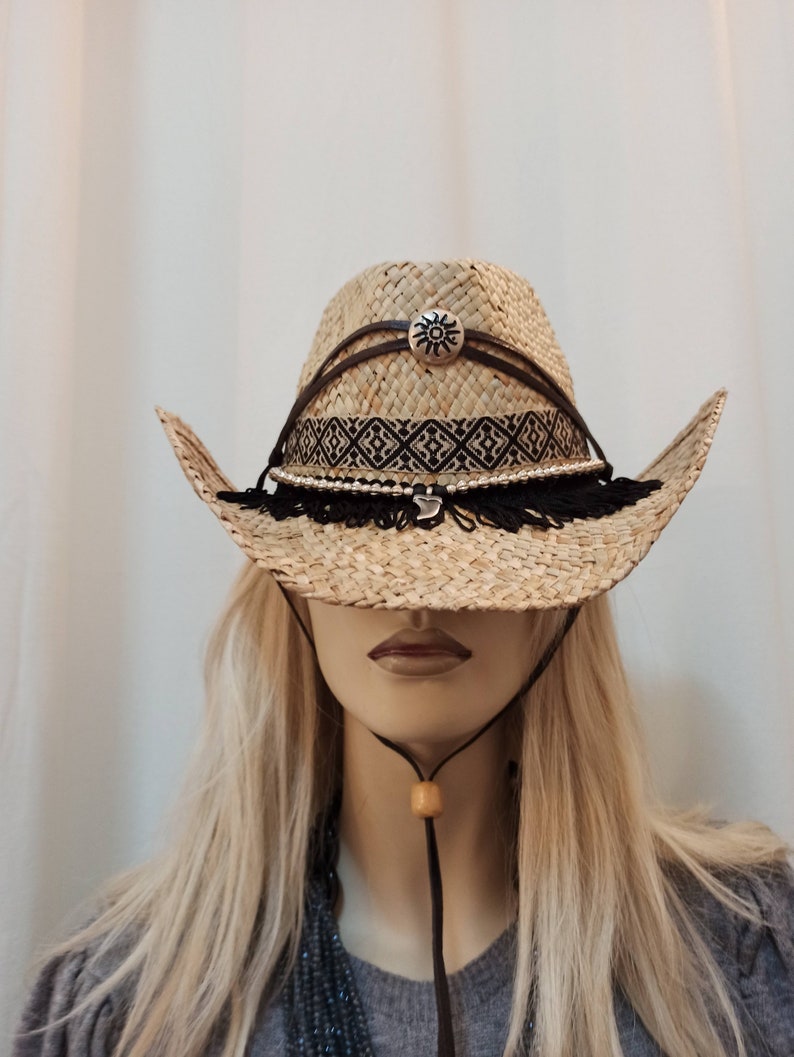 May include: A straw cowboy hat with a black and white geometric band, a silver sun charm, and black fringe trim.