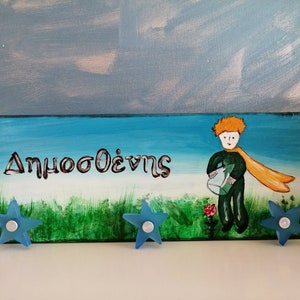 May include: A wooden sign with a blue sky and green grass background. The sign has the name "Δημοσθένης" written in white paint. A cartoon image of the Little Prince is holding a watering can. Three blue star-shaped hooks are attached to the bottom of the sign.