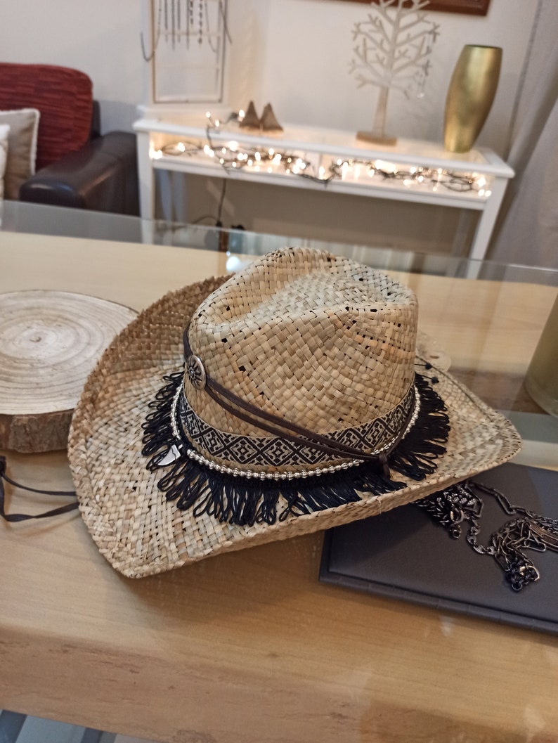 May include: A wide-brimmed straw hat with a black and white patterned band and a black fringe trim. The hat is sitting on a wooden surface.