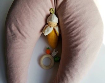 May include: A light pink, crescent-shaped nursing pillow with a small wooden ring, a carrot, and a bunny toy in front of it. A small doll with a yellow dress and a green hat is inside the pillow.