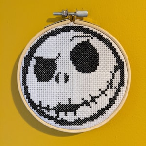 May include: A cross-stitch embroidery of Jack Skellington's face from The Nightmare Before Christmas. The design is black and white, set in a wooden embroidery hoop against a yellow background.