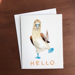 May include: A watercolor greeting card featuring a blue-footed booby bird with blue feet and a brown tail. The bird is standing on one leg and looking at the viewer. The card has the word "HELLO" written in orange letters at the bottom.