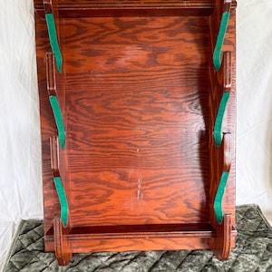 May include: A rectangular wall-mounted wooden rack with a rich brown wood grain finish. The rack features two vertical side panels, each with three curved cutouts lined with green felt, designed to hold items horizontally across the frame.