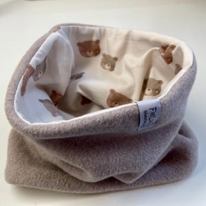 Könnte beinhalten: Ein grauer Fleece-Schlauchschal mit cremefarbenem Innenfutter. Das Innere hat ein sich wiederholendes Muster aus braunen Teddybär-Gesichtern. Ein kleines Etikett mit Text ist an der Seite angenäht. Der Schal soll den Hals warm halten.