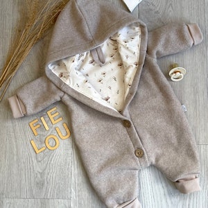 Op de afbeelding: Een beige baby-jumpsuit met capuchon en een cr&egrave;mekleurige voering met een bruin bloemenpatroon. Het kledingstuk heeft lange mouwen met geribbelde boorden, een knoopsluiting aan de voorzijde met twee zichtbare bruine knopen en de tekst FIE LOU.