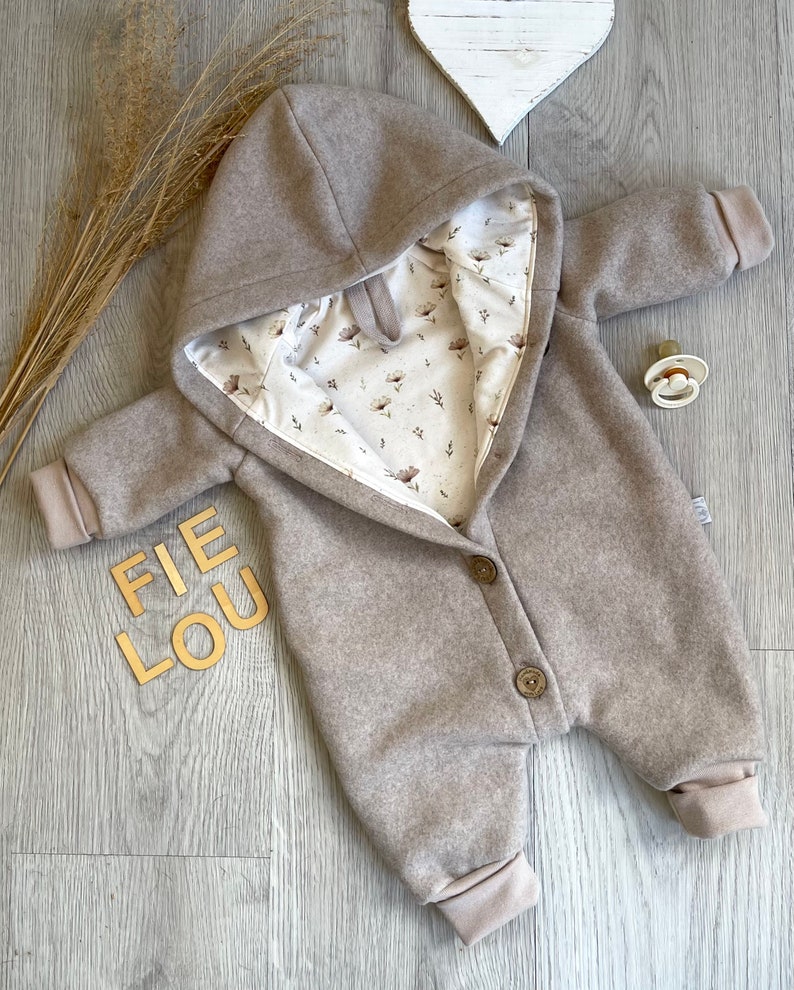 Op de afbeelding: Een beige baby-jumpsuit met capuchon en een cr&egrave;mekleurige voering met een bruin bloemenpatroon. Het kledingstuk heeft lange mouwen met geribbelde boorden, een knoopsluiting aan de voorzijde met twee zichtbare bruine knopen en de tekst FIE LOU.