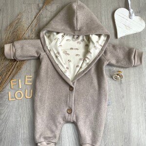 Op de afbeelding: Een beige jumpsuit met capuchon, lange mouwen en geribbelde boorden, voorzien van twee bruine knopen aan de voorzijde. De binnenvoering is cr&egrave;mekleurig met een herhalend patroon van kleine bruine egels. Een maat 68 label is zichtbaar bij de hals.
