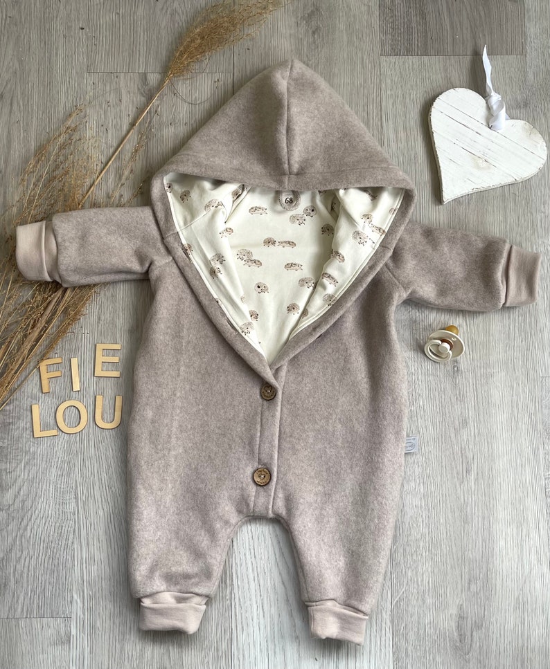 Op de afbeelding: Een beige jumpsuit met capuchon, lange mouwen en geribbelde boorden, voorzien van twee bruine knopen aan de voorzijde. De binnenvoering is cr&egrave;mekleurig met een herhalend patroon van kleine bruine egels. Een maat 68 label is zichtbaar bij de hals.