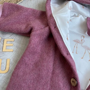Op de afbeelding: Een oudroze jas met capuchon en een witte binnenvoering met een flamingopatroon. Het kledingstuk heeft een enkele houten knoopsluiting en wordt vergezeld door een bijpassende oudroze fopspeen die bij de capuchon is geplaatst.