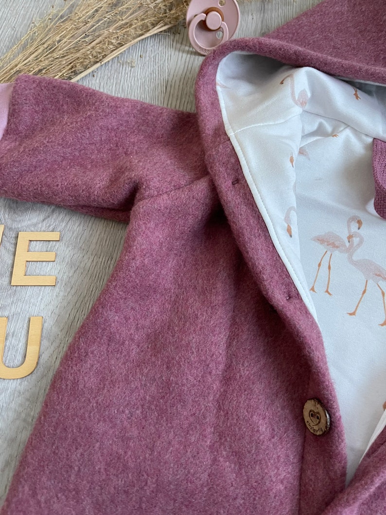 Op de afbeelding: Een oudroze jas met capuchon en een witte binnenvoering met een flamingopatroon. Het kledingstuk heeft een enkele houten knoopsluiting en wordt vergezeld door een bijpassende oudroze fopspeen die bij de capuchon is geplaatst.