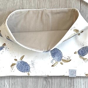 Könnte beinhalten: Eine weiße Stoff-Aufbewahrungsbox mit einem Muster aus blauen und braunen Meeresschildkröten. Die Innenseite ist einfarbig beige. Die Box ist offen und leer, mit einem kleinen Etikett in der unteren rechten Ecke.