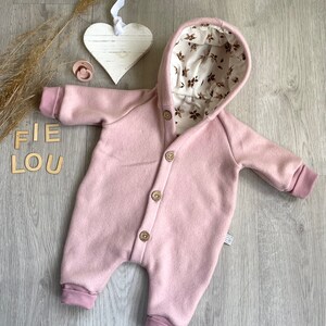 Op de afbeelding: Een oudroze jumpsuit met capuchon en een knoopsluiting aan de voorzijde met vier houten knopen. De binnenkant van de capuchon is gevoerd met een cr&egrave;mekleurige stof versierd met een bruin bloemenpatroon. Het kledingstuk heeft lange mouwen en pijpen met boorden.
