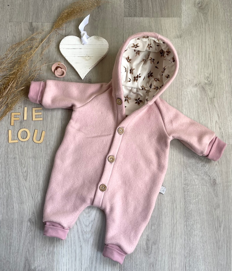 Op de afbeelding: Een oudroze jumpsuit met capuchon en een knoopsluiting aan de voorzijde met vier houten knopen. De binnenkant van de capuchon is gevoerd met een cr&egrave;mekleurige stof versierd met een bruin bloemenpatroon. Het kledingstuk heeft lange mouwen en pijpen met boorden.