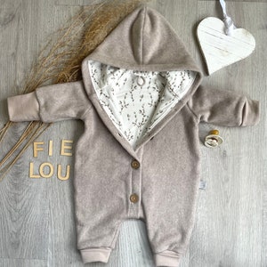 Op de afbeelding: Een beige baby-jumpsuit met capuchon, lange mouwen en geribbelde boorden. De binnenvoering is van witte stof met een zwart botanisch takjespatroon. De sluiting aan de voorzijde is voorzien van twee bruine knopen.