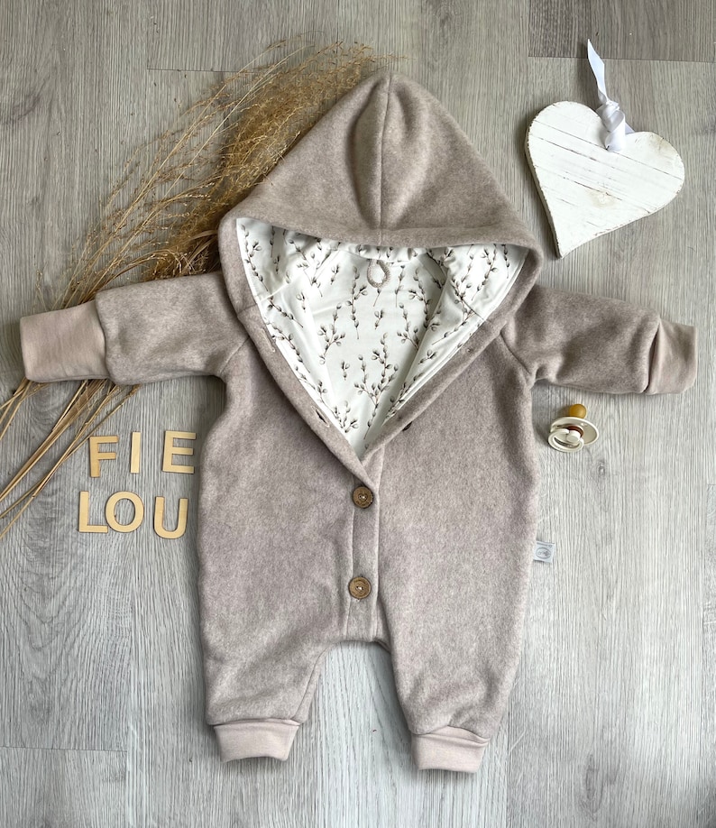 Op de afbeelding: Een beige baby-jumpsuit met capuchon, lange mouwen en geribbelde boorden. De binnenvoering is van witte stof met een zwart botanisch takjespatroon. De sluiting aan de voorzijde is voorzien van twee bruine knopen.