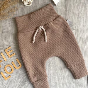 Könnte beinhalten: Braune Babyhose aus Waffelstrick mit Kordelzug und gerippten Bündchen. Die Hose hat eine lockere Passform und ist aus einem weichen, strukturierten Stoff gefertigt. Ein Schnuller und Holzbuchstaben mit der Aufschrift "IE LOU" sind ebenfalls sichtbar.