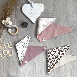 Könnte beinhalten: Eine Auswahl an Baby-Lätzchen in verschiedenen Mustern, darunter Blumenmuster, Leopardenmuster und einfarbiges Rosa. Ein Holzherz, ein gehäkeltes Bär-Rassel und ein Schnuller sind ebenfalls vorhanden. Die Lätzchen sind aus weichem Stoff und haben Druckknöpfe.